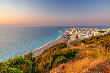 Panoramic high-angle view of Psaropoula Beach and Rhodes Town at sunset, looking towards the northern tip of Rhodes Island, Greece. Turquoise Aegean Sea, coastline, and city architecture bathed in gol © Artem