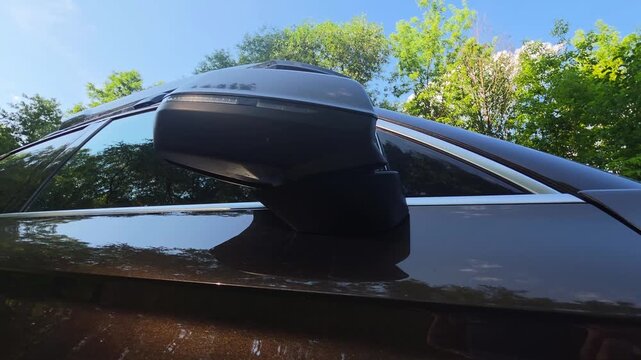 Exterior perspective of dark brown modern car side mirror, sleek design element of vehicle, blue sky with green trees reflecting on polished car body, bright sunny daytime outdoor setting
