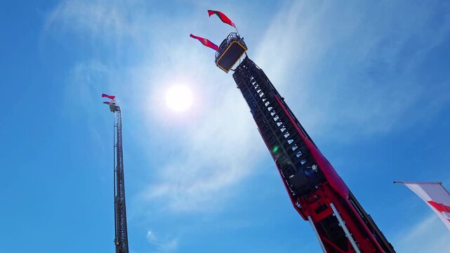 Extended red black telescopic boom fire fighting platform with multiple flags reaching high into brilliant blue sky bright sun lens flare day outdoor industrial equipment display innovation technology