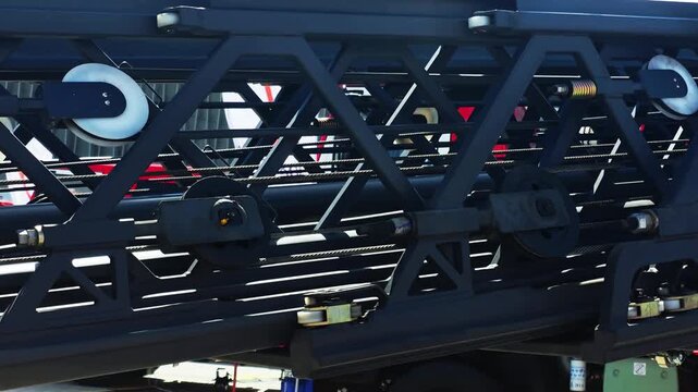 Close-up view of intricate dark gray metal framework of fire truck ladder, featuring system of cables, pulleys, and springs, demonstrating complex engineering of rescue equipment outdoors in daylight.