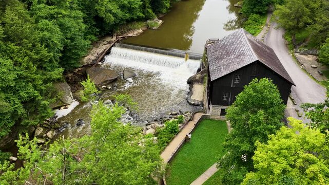 Aerial drone footage slowly approaching a historic dark-wood grist mill beside a cascading waterfall and stone dam. A red covered bridge is visible nearby, in lush greenery.