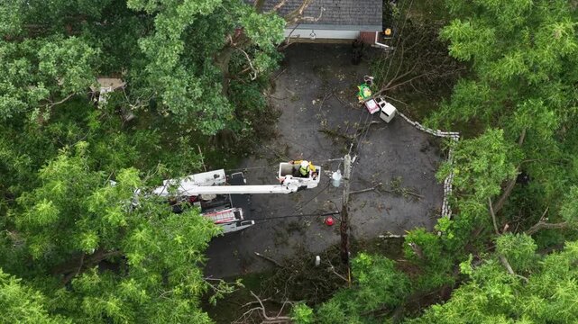 Bucket truck lineman repairs broken utility pole tangled in wires on driveway in Manheim Township, Pennsylvania, dense tree canopy surrounding storm-damaged property. Aerial drone top down shot