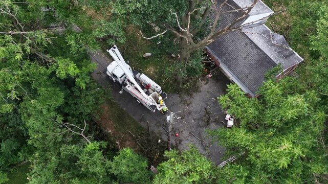 Bucket truck crew clears broken tree draped over garage roof and gravel driveway in Manheim Township, Pennsylvania, dense green yard canopy surrounding storm cleanup site. Aerial drone view.