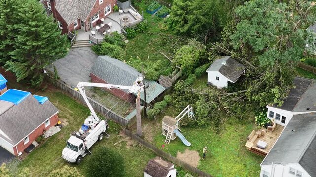 Bucket truck lineman repairs snapped utility pole beside blue tarp-covered roof in Manheim Township, Pennsylvania, fallen tree limbs scattered near backyard swing set. Aerial drone top down shot