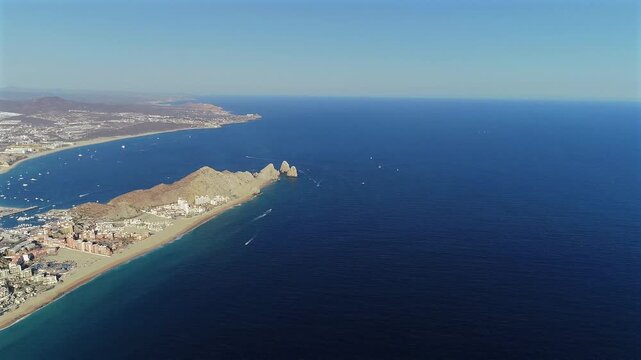 Drone flies high over the ocean starting with Lands End and the Arch in frame, panning right to reveal the coastline, the cape, boats scattered across the water, the marina, and finally the town and s