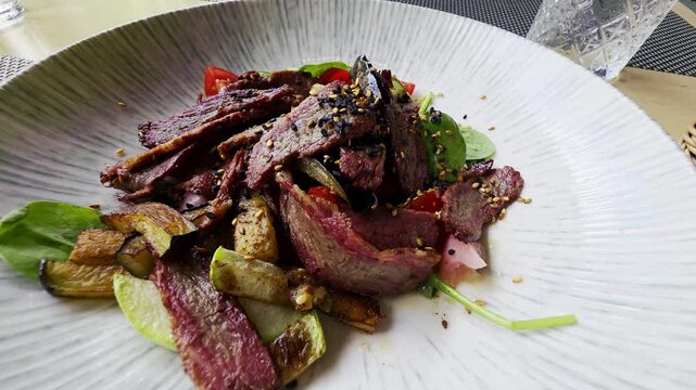 Sliced beef and roasted vegetable salad with fresh greens, tomatoes, and sesame seeds on decorative white plate. Healthy meal showcasing rich protein and vibrant ingredients.