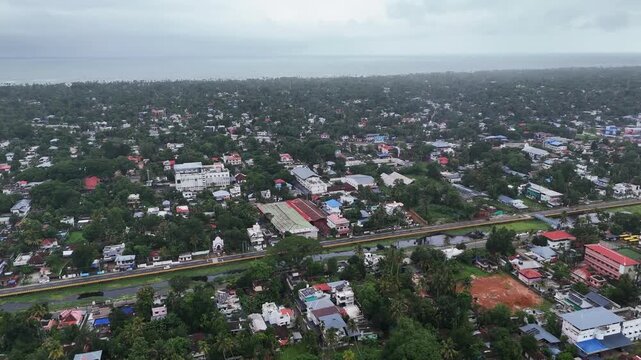 Cinematic drone footage of Alappuzha featuring lush neighborhoods, a scenic canal, and the Arabian Sea on the horizon. A stunning aerial view blending Kerala's tropical beauty with its vibrant urban