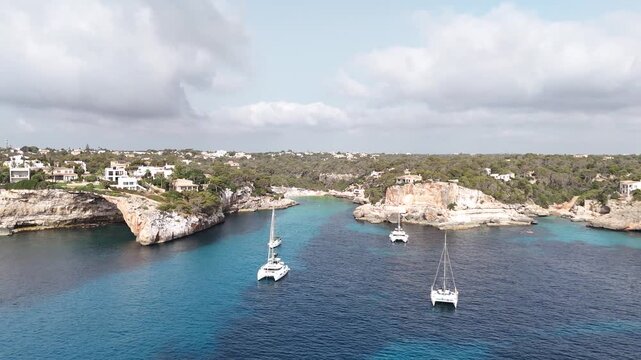 A sweeping pan shot showing boats and yachts anchored in the water at Cala Lombardes, Mallorca, Spain. The shot shows the boats and the landscape in the background.