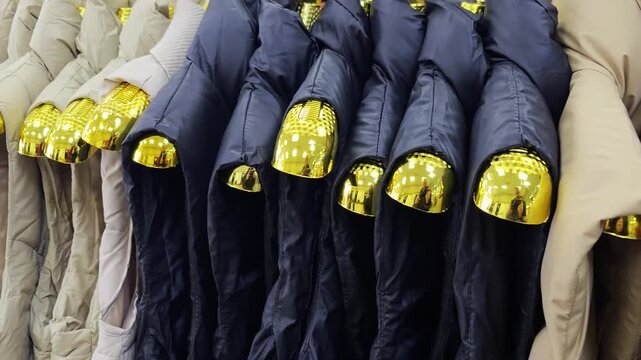 Row of colorful winter puffer jackets on golden hangers inside clothing store, showcasing various warm outerwear for cold weather conditions and fashion retail