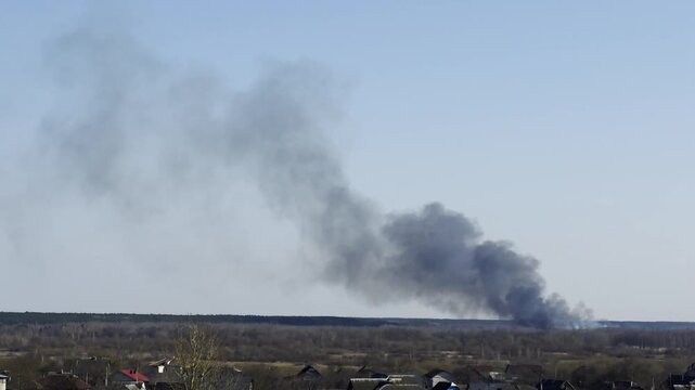 Thick black smoke rising from fire in rural landscape with houses below and clear blue sky overhead, visible environmental impact of burning incident