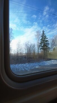 View through train window showing passing landscape ground covered with patches of melting snow, sparse trees. Early spring nature under bright blue sky with white clouds. Winter. Vertical shot