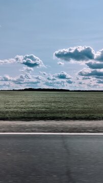 View through vehicle window with car driving along asphalt road adjacent to expansive green meadow under blue sky with fluffy white clouds overhead. Open field, cumulus clouds. Vertical shot