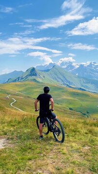 Male cyclist  with mountain electric bike on grassy hill looking at majestic alpine peaks under blue sky