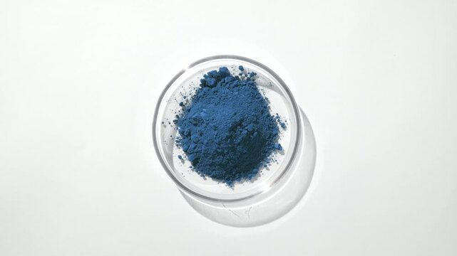 Top view frame of a pile of blue powder contained in a glass petri dish on a light grey backdrop lit with orbiting light and casts moving shadow
