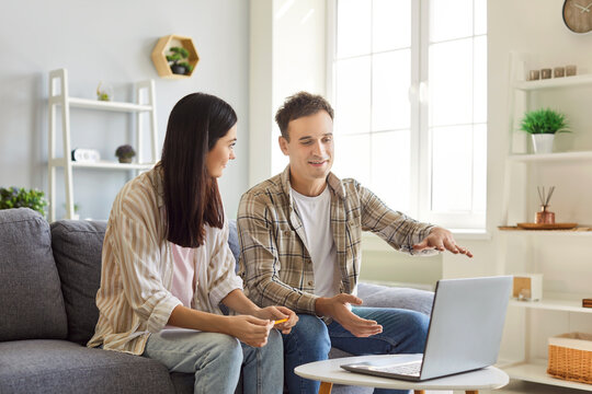 Portrait of a young smiling married couple sitting on sofa at home looking at laptop discussing ideas about interior design. Man and woman planning repair. Renovation and moving concept.