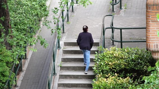 Mujer coja y piernas arqueadas y torcidas se&ntilde;ora sudamericana subiendo pelda&ntilde;os de escalera en calle con plantas, cuesta y barandilla 
