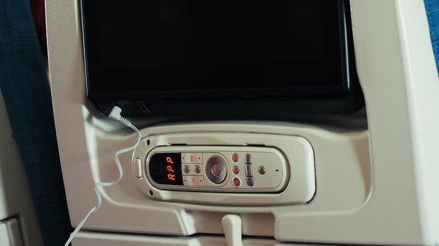 Seatback entertainment screen and passenger control panel inside a commercial airplane cabin. Close-up of an aircraft seat with in-flight entertainment system, headphone connection and remote controls