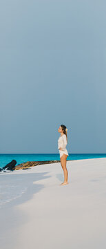 Young woman in white summer outfit enjoying sunny day on tropical sandy beach with turquoise ocean water