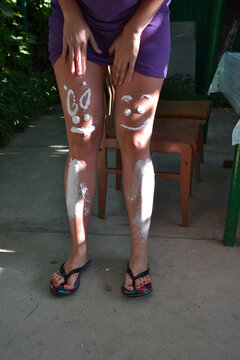 Woman applying sunscreen creatively on her legs with funny smiley faces before sunbathing outdoors. Summer skincare, UV protection, vacation, beach preparation, healthy lifestyle and sun safety