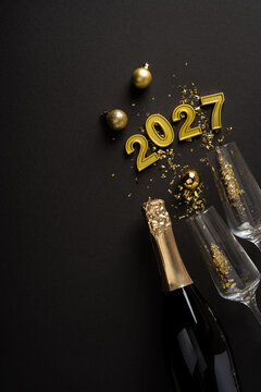 Happy New Year 2027 celebration with gold champagne bottle, glass and festive confetti on black background on vertical black tabletop