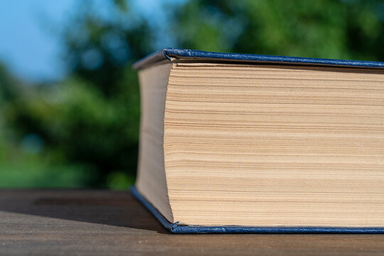 Closeup of thick vintage book outdoors. Side view macro shot of thick hardcover book with yellowed paper pages resting on wooden surface against blurred green garden background