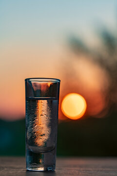 Cold shot glass with clear alcohol on table. Closeup of sweaty condensation covered shot glass filled with strong transparent alcohol or vodka against beautiful warm blurred sunset sky background