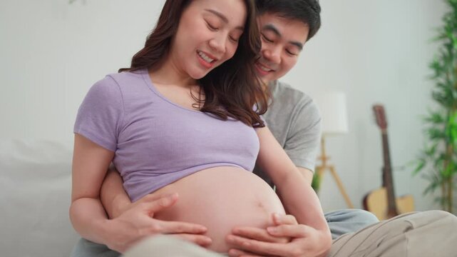 Asian husband and pregnant wife sit together on cozy bed in bedroom. 