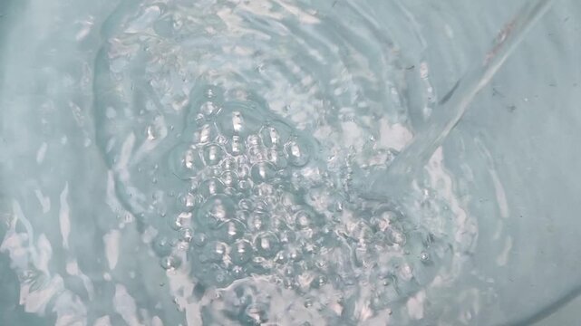 Overhead top view of clean clear water being poured into a light blue bucket creating a swirling vortex, ripples and air bubbles. Abstract water background texture, slow motion water splash