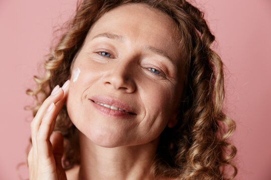 A middle aged woman with natural skin applies cream to her face, smiling in a pink studio. A candid beauty moment centered on selfcare and healthy aging with calm and natural glow.