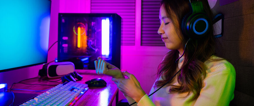 Young Asian woman gamer wearing headphones playing video game on modern gaming pc computer have long time she wrist pain, female broadcast streaming plays online home games
