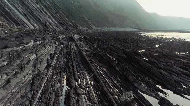A breathtaking cinematic low-angle tracking view capturing the dramatic, multi-layered vertical rock strata of the Flysch de Zumaia along the rugged Basque Country coastline.