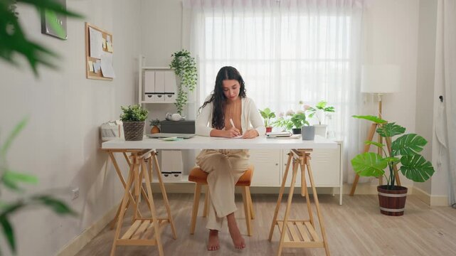 Young Hispanic woman writing while sitting at table in living room at home. 