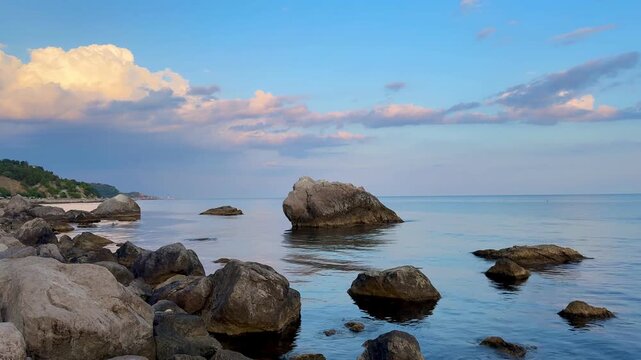 Calm sea and coastal boulders at golden hour, blue sky with clouds
