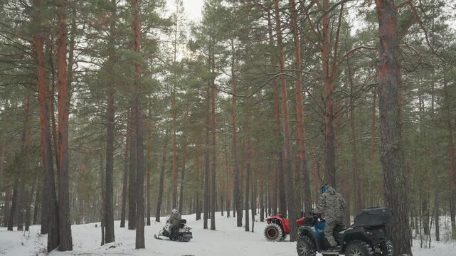 Snow pine atv convoy gathers among pines, several riders on colorful quads pause on snowy trail, red and blue machines line up, helmeted drivers check maps and chat, scattered tire tracks and snow