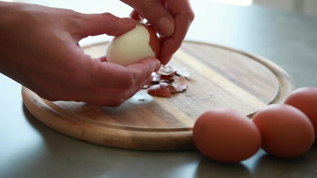 Close Up of Female Hands Peeling Hard Boiled Brown Egg on Wooden Cutting Board