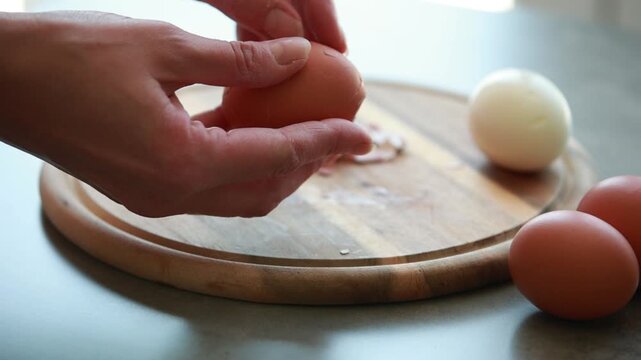 Close Up of Female Hands Peeling Hard Boiled Brown Egg on Wooden Cutting Board
