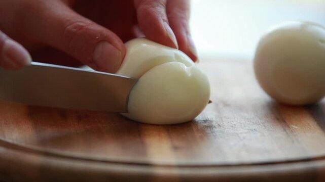 Female Hands Slicing Hard Boiled Egg with Knife on Wooden Cutting Board