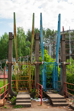 View of an old, weathered amusement park ride with yellow and blue metal cages and concrete supports, set against trees and roller coaster tracks in Elektrenai, Vilnius County, Lithuania.