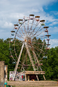 View of an abandoned ferris wheel with graffiti-covered base surrounded by lush green trees under a blue sky in Elektrenai, Vilnius County, Lithuania.