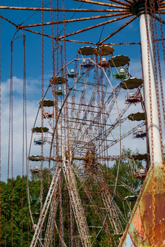 View of an old, rusty Ferris wheel in an amusement park, with swing ride chains in the foreground and green trees under a blue sky in Elektrenai, Vilnius County, Lithuania.