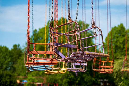 View of rusted, colorful chain swing ride seats in an amusement park with lush green trees in the background under a clear blue sky in Elektrenai, Vilnius County, Lithuania.