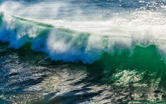 A vibrant green ocean wave breaks forcefully, sending a plume of white spray into the air under the bright sunlight, creating a dynamic and energetic natural scene. The deep blue water reflects the