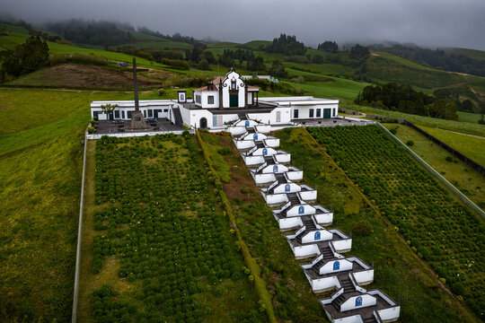 Aerial view of Chapel of Nossa Senhora da Paz with its iconic zigzag staircase, surrounded by lush green hills and agricultural fields in Vila Franca Do Campo, Acores, Portugal.