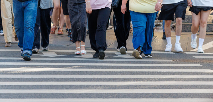 Pedestrians cross street at zebra crossing. Diverse people walk in different attire. Legs and feet are the main focus. Crosswalk lines mark the crossing path.