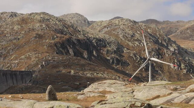 Wind turbine and dam wall at Gotthard Pass, Switzerland. Static shot of renewable energy in the Swiss Alps, rotor turning above autumn slopes with hydropower dam and rocky terrain