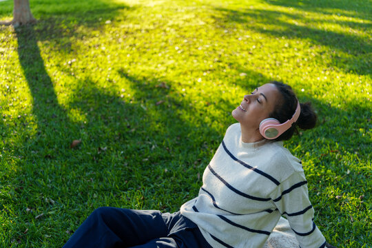 Woman in her 40s enjoys leisure time, sitting on park grass while listening to music through pink headphones