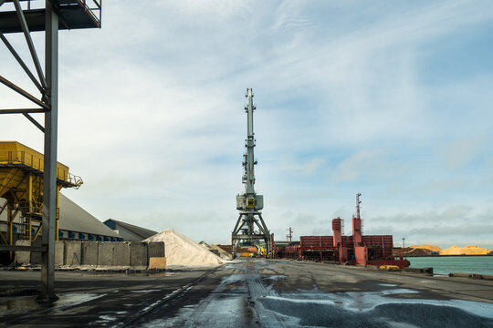 View of the Port of Ventspils featuring a large industrial crane, piles of bulk material, and harbor infrastructure in Ventspils, Ventspils Municipalit, Latvia.