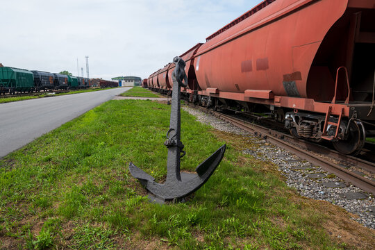 View of a large black anchor displayed on the grass beside a railway track with red freight train cars at the Port of Ventspils, Ventspils, Ventspils Municipalit, Latvia.