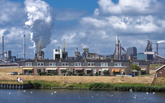 View of the Tata Steel industrial plant with smoking chimneys behind a row of traditional Dutch houses and a canal with swans in Velsen-Noord, Noord-Holland, Netherlands.