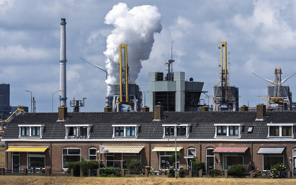 View of Tata Steel industrial plant with smoke stacks and cranes rising behind a row of residential houses in Velsen-Noord, Noord-Holland, Netherlands.
